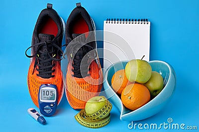 Orange sneakers, notepad, glucometer, measuring tape and fruit in a heart-shaped bowl on a blue background Stock Photo