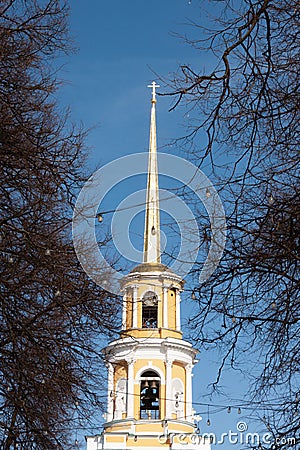 A orange and yellow golden orthodox church with a long pillar and a cross on top. A bell can be seen inside. The building is Stock Photo