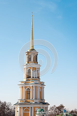 A orange and yellow golden orthodox church with a long pillar and a cross on top. A bell can be seen inside Stock Photo