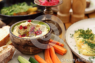 Organic humus in a bowl, decorated with pomegranate seeds, served with vegetable snacks, on a plate, on a wooden table. Stock Photo