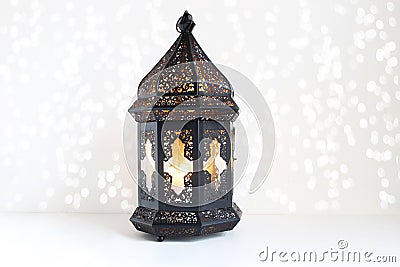 Ornamental dark Moroccan, Arabic lantern on the white table. Burning candle, glittering bokeh lights. Greeting card for Stock Photo