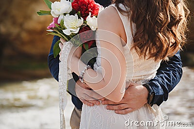 Outdoor beach wedding ceremony near the ocean, close up of embrace of stylish couple with wedding bouquet, man is hugging bride Stock Photo