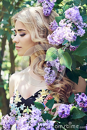 Outdoor fashion beautiful young woman surrounded by lilac flowers summer. Spring blossom lilac bush. Portrait of a girl blond Stock Photo