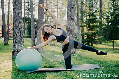 Outdoor shot of active brunette woman in sportswear poses on yoga mat, does stretching exercises with gymnsatic ball, poses in Stock Photo
