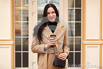 Outdoor shot of cheerful adorable young lady with red lips posing over glass door, looking directly at camera, holding papercup of Stock Photo