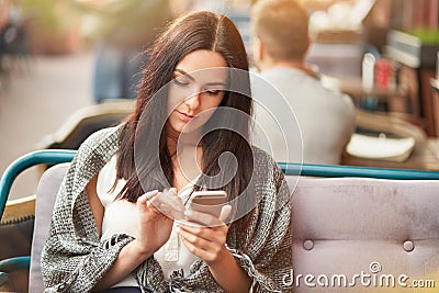 Outdoor shot of concentrated serious young female holds mobile phone in hand, types message to boyfriend, surfes social network or Stock Photo