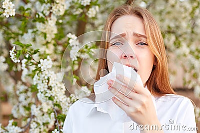 Outdoor shot of discontent young girl has seasonal allergy, uses tissue, poses over blooming tree, has rhinitis and sneezing, Stock Photo