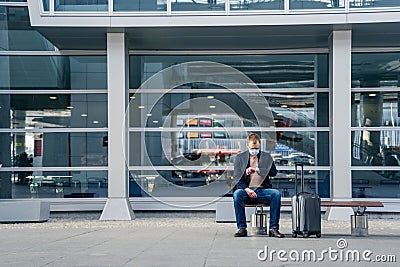 Outdoor shot of male passenger waits for transport on bench, poses at station, looks at watch, suitcase on wheels near, wears Stock Photo