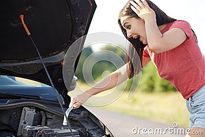 Outdoor shot of puzzled pretty female stares at opened car hood with surprised expression, tries to repair engine by herself, has Stock Photo