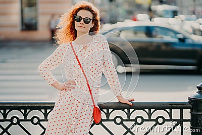 Outdoor shot of satisfied redhead woman keeps one hand on waist, other on street hence, poses over blurred urban setting, wears Stock Photo