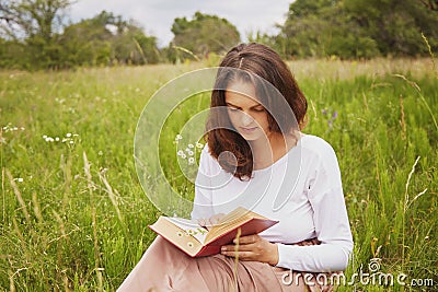 Outdoor shot of serious lovely young woman enjoys summer weather, sits on green field, reads interesting book with great interest, Stock Photo