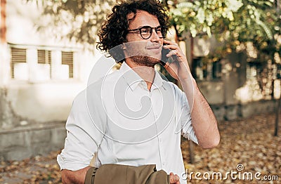 Outdoor shot of a smart young man talking on mobile phone in the street. Handsome male with curly hair in a white shirt and eyegla Stock Photo