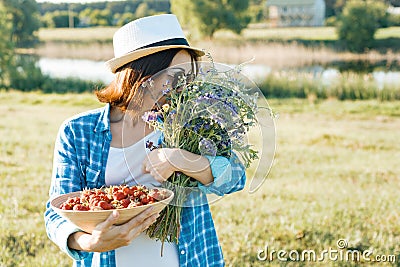 Outdoor summer portrait of adult woman with strawberries, bouquet of wildflowers, straw hat and sunglasses. Nature background, Stock Photo
