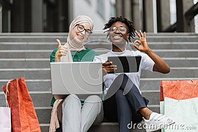 African and Muslim women sitting on the steps of mall with laptop and tablet and shopping online. Stock Photo