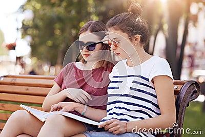 Outdoor view of serious concentrated two schoolgirls reads textbook attentively, try to learm material for lesson, enjoy fresh air Stock Photo
