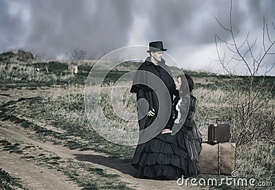 Outdoors portrait of a victorian lady in black sitting on the road with her luggage and gentleman standing nearby Stock Photo