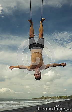 Outdoors portrait of young attractive and athletic man practicing aero yoga workoutat the beach hanging from rope swiing above the Stock Photo