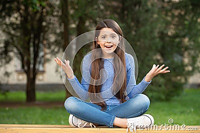 Outraged girl teenager shrugging shoulders sitting legs crossed on bench outdoors, outrage Stock Photo