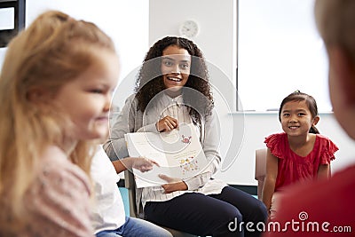 Over shoulder view of female teacher showing a picture in a book to a group of kindergarten children sitting on chairs in a classr Stock Photo