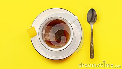 Overhead shot - porcelain cup with hot water, teabag just put in, staining the liquid, silver spoon next to it on yellow board Stock Photo