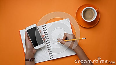 Overhead shot of stylish holding mobile phone and making note in notebook on orange background. Stock Photo