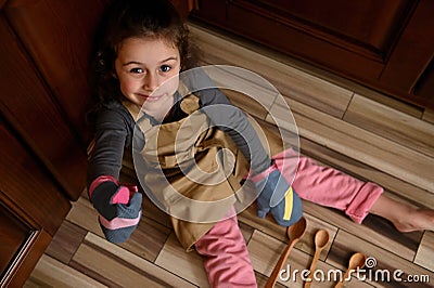 Little girl baker confectioner, chef pastry in chef apron and mittens shows thumbs up, sitting barefoot on kitchen floor Stock Photo