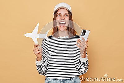 Overjoyed happy cheerful woman wearing baseball cap and striped shirt holding white paper plane and phone isolated over beige Stock Photo