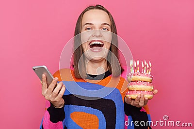 Overjoyed woman holding delicious appetizing doughnuts with burning candles wearing casual jumper using cell phone, standing Stock Photo