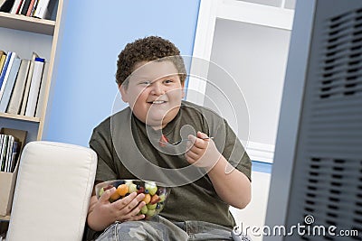 Overweight Boy Eating Bowl Of Fruit In Front Of TV Stock Photo