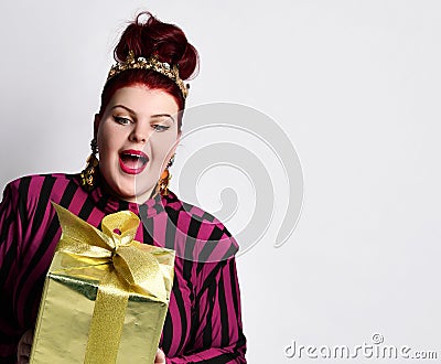 Overweight ginger female in striped dress, crown and earrings. Holding golden gift box, excited, posing isolated on white Stock Photo
