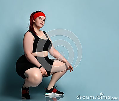 Overweight ginger model in red headband, black top, shorts, sneakers. Squatting posing sideways against blue studio background Stock Photo