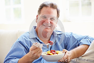 Overweight Man Sitting On Sofa Eating Bowl Of Fresh Fruit Stock Photo