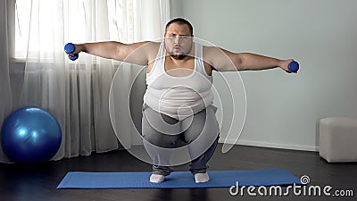 Overweight man squatting and lifting dumbbells on mat, full body training home Stock Photo
