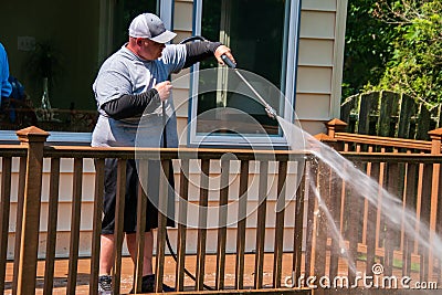 Overweight man wearing white baseball cap power washing the railing of a brown wooden deck in a backyard. Editorial Stock Photo