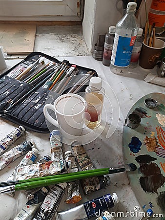 Paintbrushes, artist palette, pencils, coffee cup, watercolor and oil paints on desk in painter studio. Stock Photo