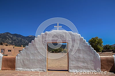 Painted doorway topped by cross with mountains and blue sky and man carrying bucket viewed through it at Taos Pueblo in New Mexico Stock Photo