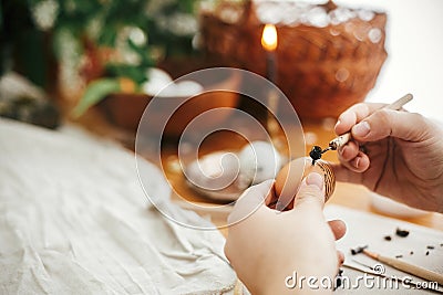 Painting Easter egg with hot wax on background of rustic wooden table with candle, basket, greenery. Easter egg with modern Stock Photo