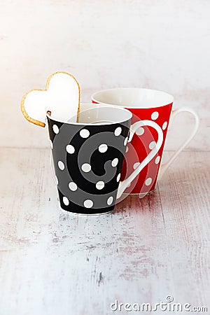 A pair of black and red polka dotted coffee cups with a heart shaped homemade cookie on the edge over white wood background. Stock Photo
