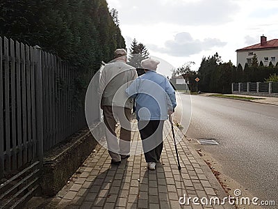 A pair of elderly people walk along the sidewalk along the road holding hands. Grandfather and grandmother on a walk in a Editorial Stock Photo