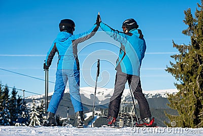 Pair giving high five to each other, smiling, standing with skis on mountain top at a winter resort with ski lifts Stock Photo