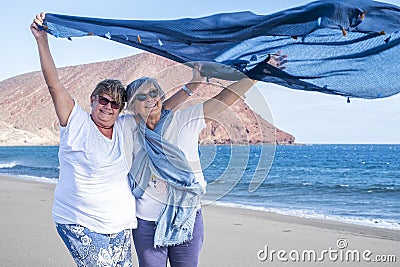 A pair of smiling and cheerful sisters enjoying the winter sea on a windy day holding a scarf in the sea breeze. Active elderly Stock Photo