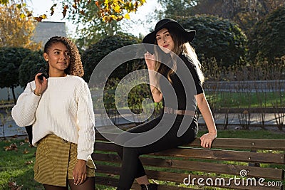 Pair of young beautiful mexican generation y girls. The girl is sitting on the back of the bench, a girlfriend is standing next to Stock Photo