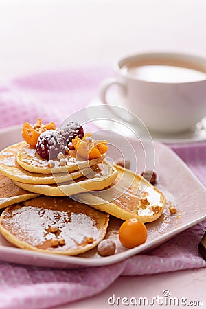 Pancakes with raspberries, physalis, hazelnut and honey on pink plate, sprinkled with powdered sugar, with fork and cup of coffee Stock Photo