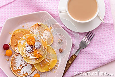 Pancakes with raspberries, physalis and honey on pink plate, sprinkled with powdered sugar, with fork and cup of tea or coffee on Stock Photo