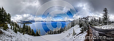 Panorama of Crater Lake with a road on the right, deep blue water, snow covered pine trees and dramatic clouds Stock Photo