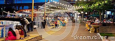 Panorama view blurry people at waterfront boardwalk string lights, beach chairs, patio restaurant dining tables blue hour with Editorial Stock Photo