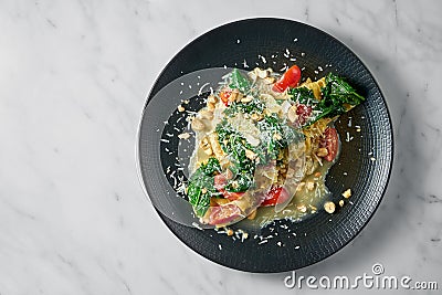 Pappardelle pasta with parsnip, cream sauce, blanched greens, tomatoes and nuts on a black plate on a white marble background Stock Photo