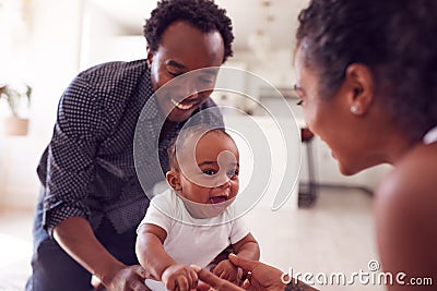 Parents Encouraging Smiling Baby Daughter To Take First Steps And Walk At Home Stock Photo