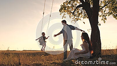 Parents swing a happy child high up on a swing at sunset, a cheerful family in the glare of the sun plays with their Stock Photo