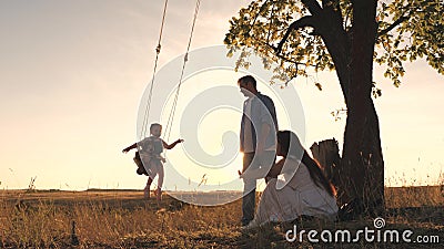 Parents swing a happy child high up on a swing at sunset, a cheerful family in the glare of the sun plays with their Stock Photo